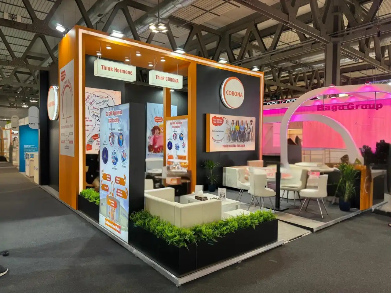 Corner trade show booth for Corona with orange and black panels, illuminated signs, white lounge seating, planters, and promotional displays in an expo venue.