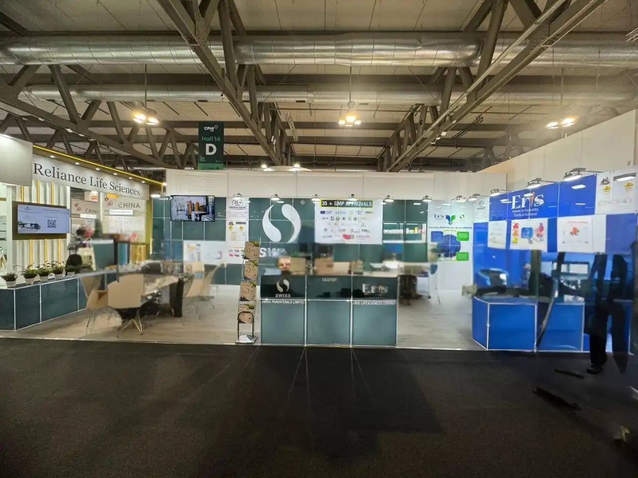 Wide pharmaceutical expo aisle featuring Reliance Life Sciences, Swiss, and Eris branded booths with glass counters, display screens, and meeting tables.