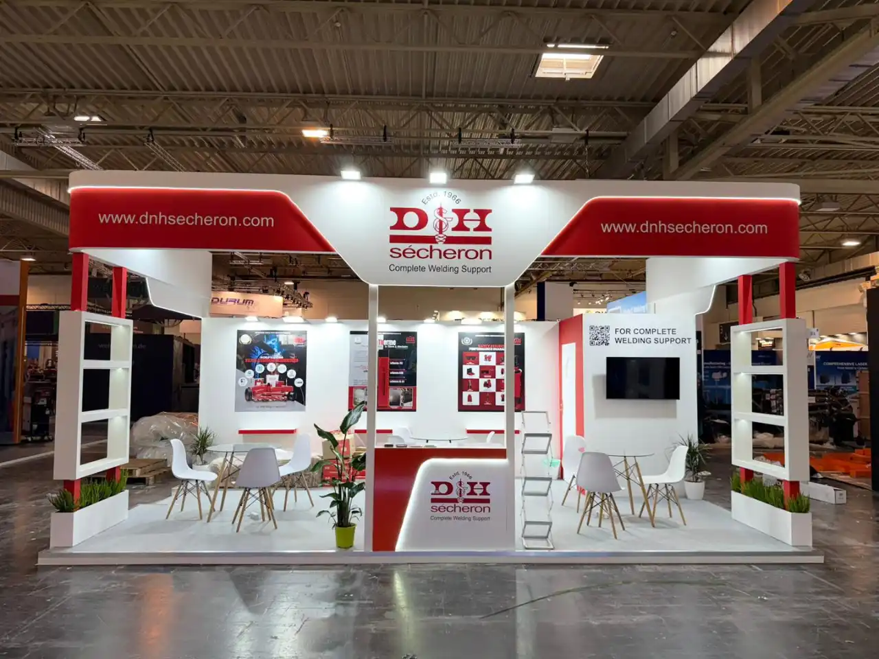 Red and white DH Sécheron exhibition booth with bold branding, central counter, display panels, seating tables, and open structure in convention center.