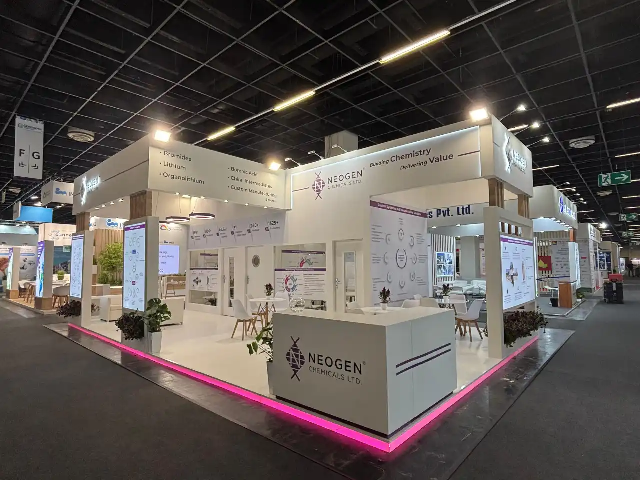 Spacious Neogen Chemicals Ltd exhibition booth with white minimalist design, pink floor lighting, digital screens, counters, plants, and meeting tables.